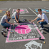 Three students painting a 'Class of 2027' design on a parking lot with a pink and white volleyball graphic.