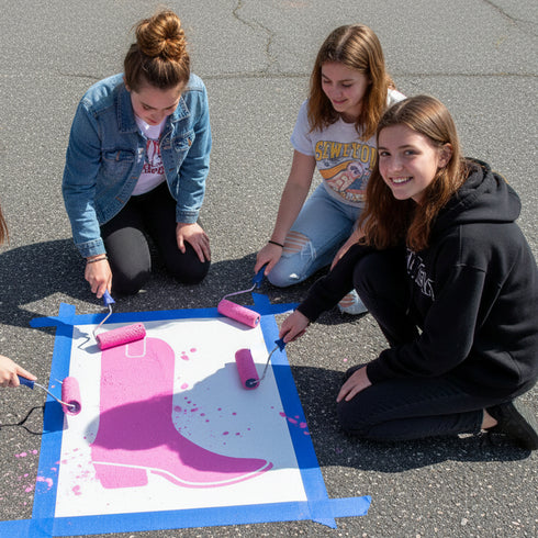 Three people painting a pink boot design on a surface outdoors.