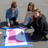 Three people painting a pink boot design on a surface outdoors.